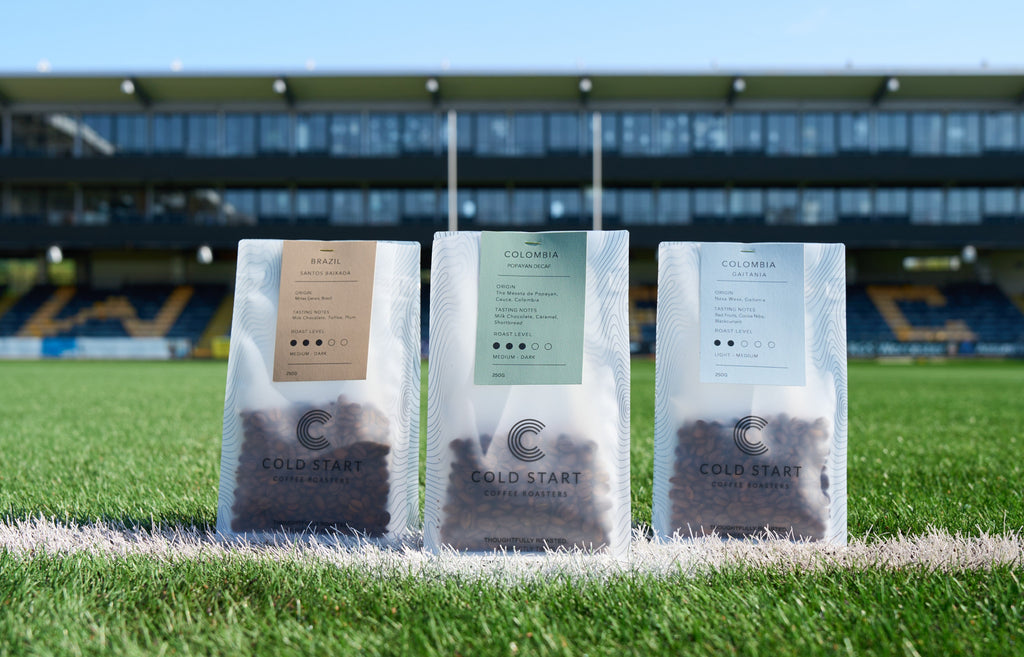 Three stand-up pouches of Cold Start Coffee Roastery coffee beans are lined up on a white field line with a green grass field and a stadium in the background under a blue sky.
